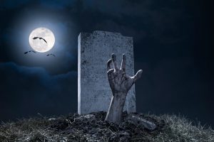 A zombie hand bursting through the grave in a graveyard on Halloween night under a full moon with vampire bats.